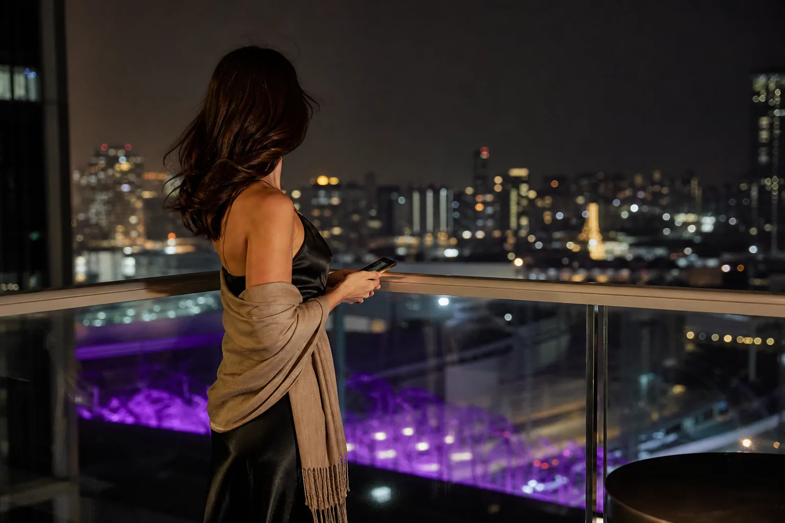 Adult creator in a long satin evening dress on a hotel balcony at night with city lights behind her, face turned away